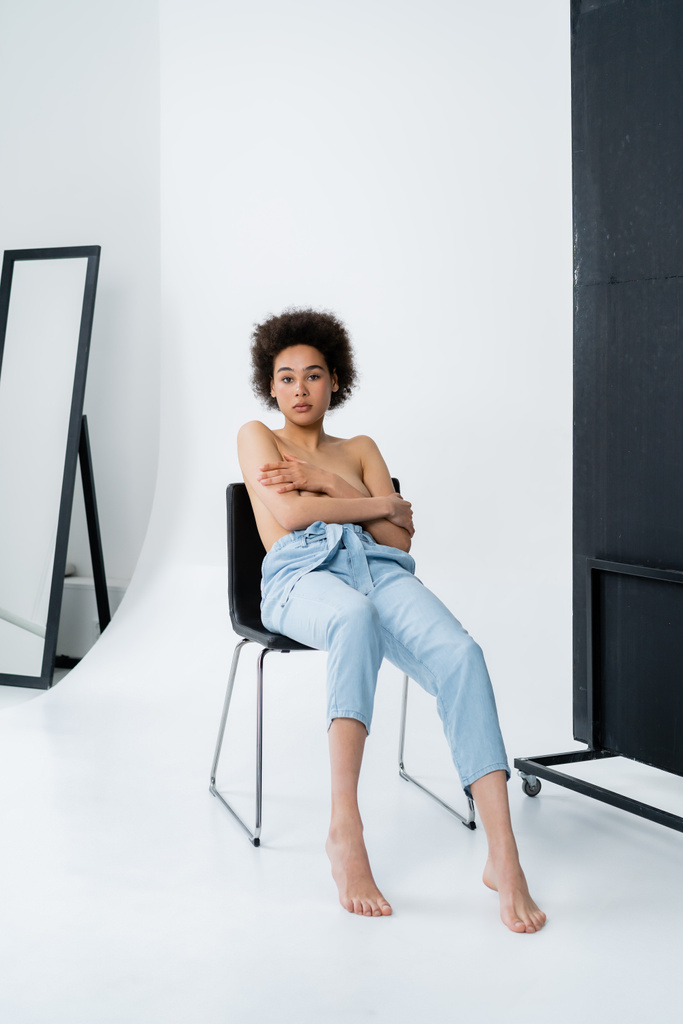 Безсоромна афроамериканська жінка в штанах сидячи на стільці на сірому фоні.  - Фото, зображення