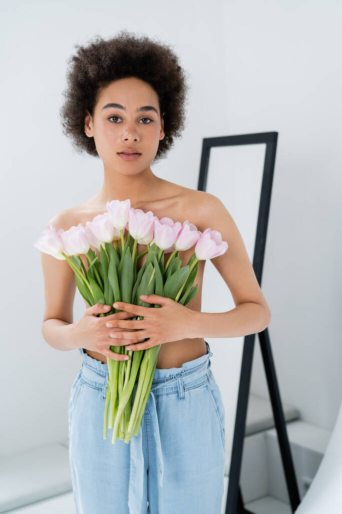 Shirtlose afrikanisch-amerikanische Frau hält Tulpen in Brustnähe auf grauem Hintergrund  - Foto, Bild