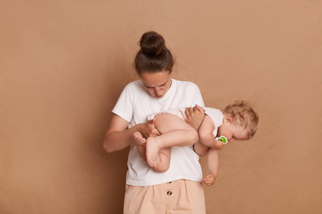 Photo de maman regardant dans la couche puante de sa petite fille, tenant un enfant en bas âge et faisant une grimace pour le dégoût, debout isolé sur fond brun. - Photo, image