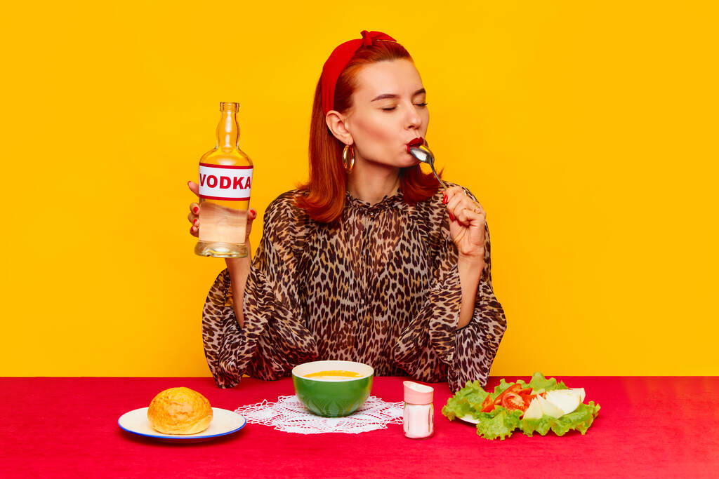 Elegante pelirroja sentada a la mesa sobre fondo amarillo y cenando, comiendo sopa, ensalada y bebiendo vodka. Fotografía de arte pop de comida. Colores complementarios. Copiar espacio para anuncios, texto - Foto, Imagen