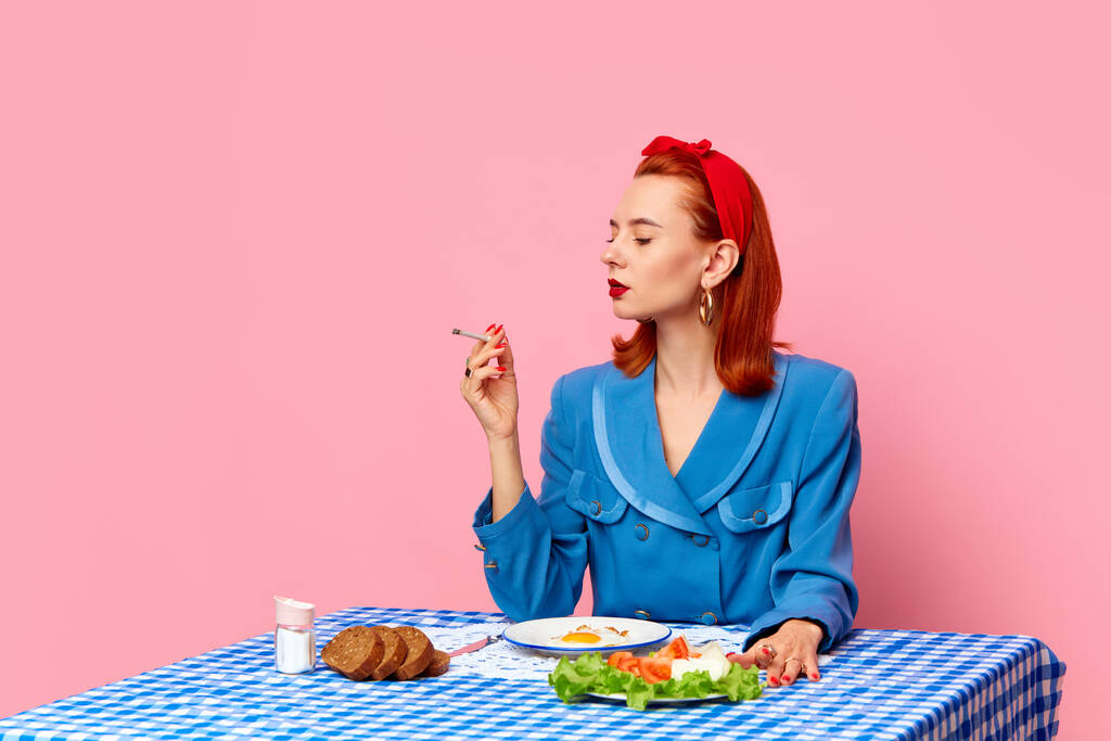 Fotografia di food pop art. Elegante donna rossa seduta a tavola, che fuma e fa colazione all'inglese con uova fritte sullo sfondo dello studio rosa. Colori complementari. Copia spazio per annuncio, testo - Foto, immagini
