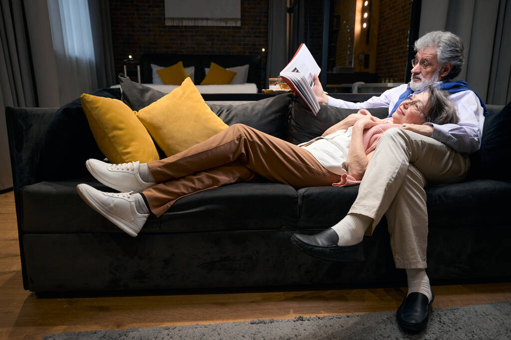 Літній чоловік і леді відпочивають на дивані в готелі, чоловік тримає книгу і читає своїй дружині
 - Фото, зображення