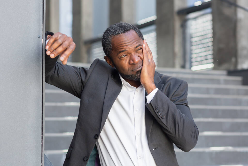 Mal di denti per strada. Un anziano afroamericano in giacca e cravatta sta fuori da un ufficio e gli tiene la guancia, soffrendo di mal di denti.. - Foto, immagini