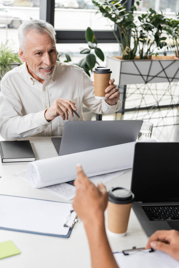 Pozytywny biznesmen w średnim wieku trzymający kawę w pobliżu laptopów i afrykańskiego kolegi w biurze  - Zdjęcie, obraz