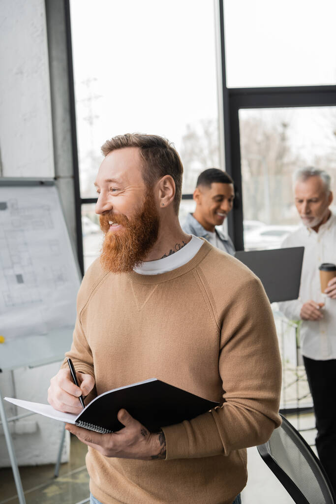 Positive bearded businessman holding notebook while working with blurred interracial colleagues in office  - Photo, Image