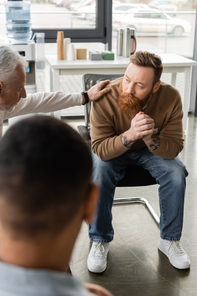 Чоловік середнього віку заспокоює татуйовану людину з алкогольною залежністю під час терапії в реабілітаційному центрі
  - Фото, зображення