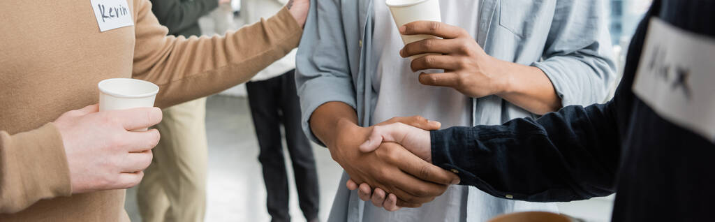 Vista recortada de personas multiétnicas con adicción al alcohol sosteniendo vasos de papel y estrechando las manos en el centro de rehabilitación, pancarta  - Foto, imagen