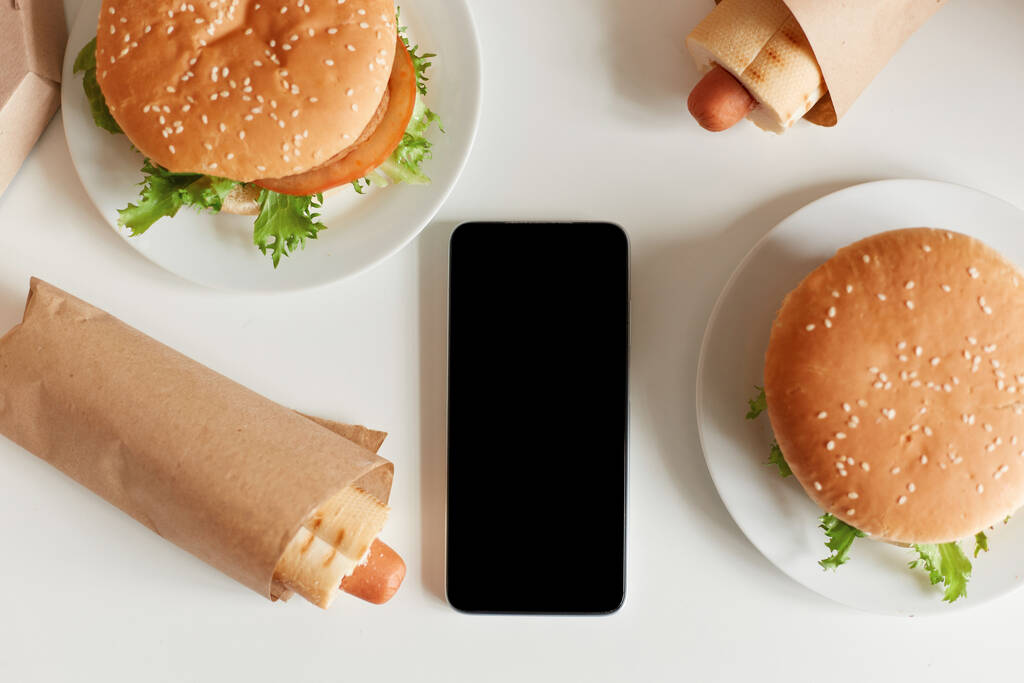 Widok z góry na niezdrowe jedzenie na stole, smaczna kiełbasa w cieście i duże hamburgery, smartfon z czarnym, pustym ekranem, przestrzeń na reklamę lub tekst promocyjny. - Zdjęcie, obraz