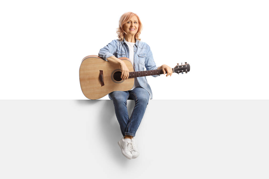 Full length portrait of a mature woman sitting on a panel with an acoustic guitar isolated on white background - Foto, Bild
