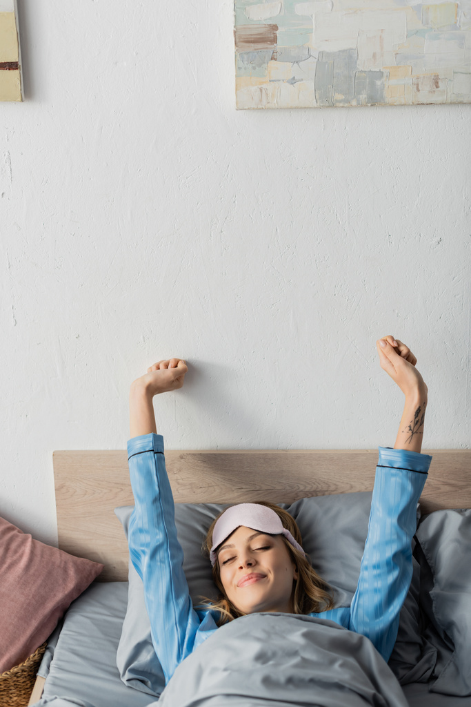 татуированная женщина в маске для сна и ночной одежде, растягивающаяся, лежа в постели  - Фото, изображение