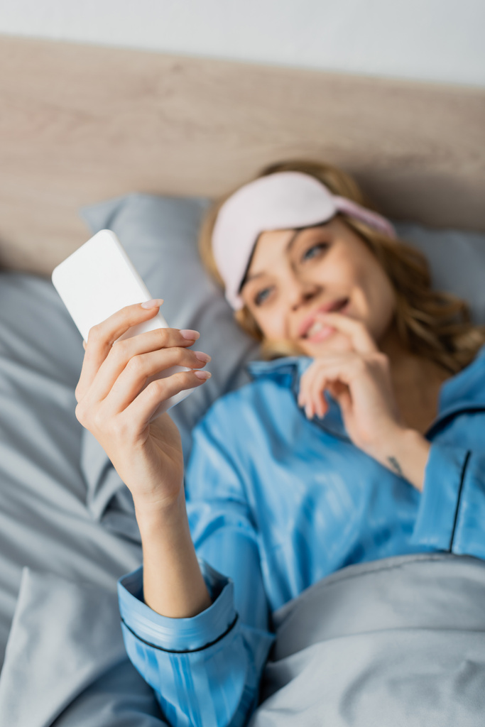 verschwommene und fröhliche Frau in Schlafmaske und blauem Pyjama mit Smartphone im Bett  - Foto, Bild