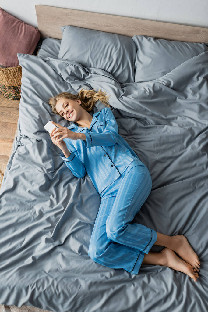 horní pohled na veselou ženu v modrém pyžamu pomocí smartphone v posteli  - Fotografie, Obrázek