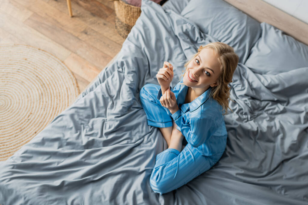 Вид сверху положительной женщины в синей пижаме, смотрящей в камеру сидя на кровати  - Фото, изображение