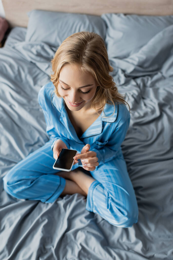 Draufsicht einer lächelnden Frau im blauen Pyjama mit Smartphone mit leerem Bildschirm, während sie im Bett sitzt  - Foto, Bild