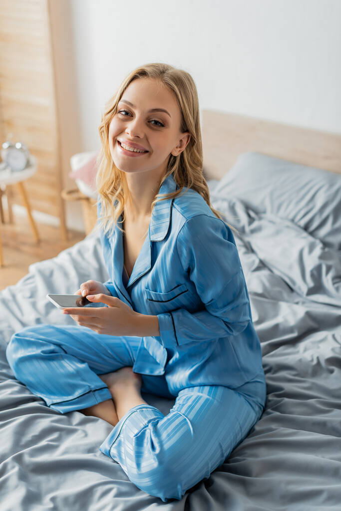 femme positive en pyjama bleu tenant smartphone avec écran blanc  - Photo, image