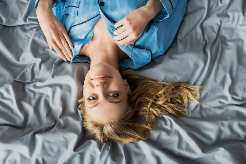 Draufsicht einer glücklichen Frau im blauen Seidenpyjama, die auf dem Bett liegt und in die Kamera blickt  - Foto, Bild
