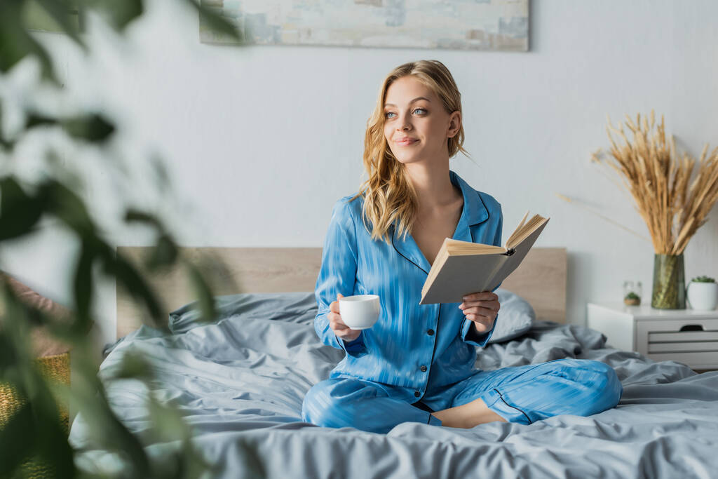 csinos fiatal nő kék selyem hálóing gazdaság könyv és csésze kávé a modern hálószobában  - Fotó, kép