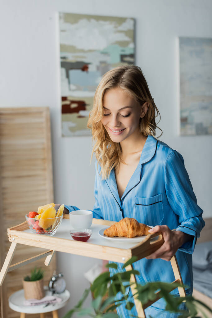 gelukkig vrouw in blauw pyjama kijken naar lade met ontbijt in slaapkamer  - Foto, afbeelding