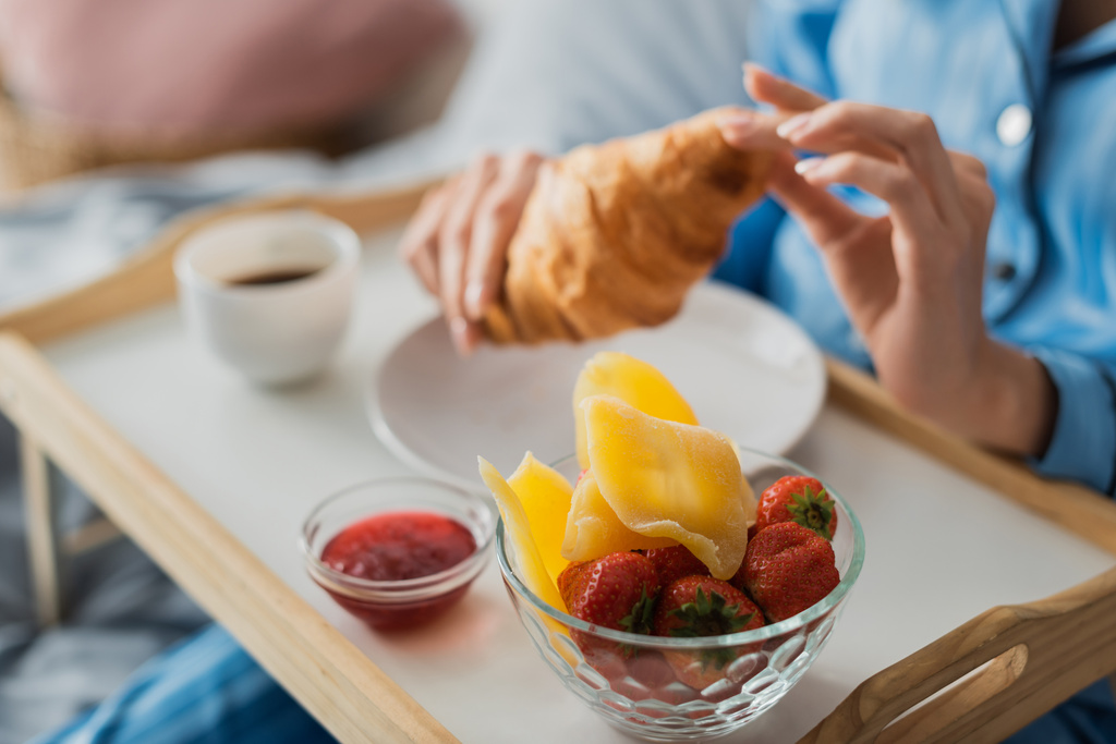 kivágott kilátás nő gazdaság friss croissant közel tálca lekvárral és szárított mangó, miközben reggeli az ágyban  - Fotó, kép