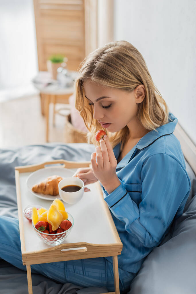 blonde junge Frau im Pyjama hält Tasse Kaffee und frische Erdbeere in der Nähe Tablett beim Frühstück im Bett  - Foto, Bild