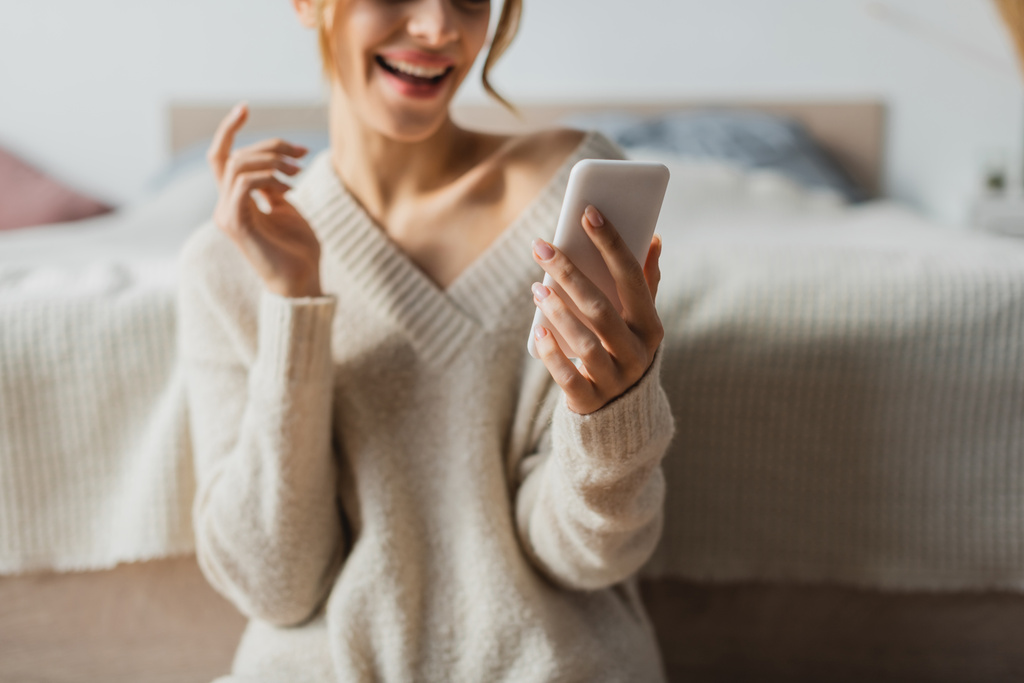 abgeschnittene Ansicht einer glücklichen Frau im Strickpullover mit Smartphone im modernen Schlafzimmer  - Foto, Bild