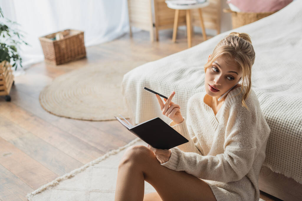 junge blonde Frau im Pullover spricht mit Smartphone und hält Stift in der Nähe von Notizbuch im Schlafzimmer  - Foto, Bild