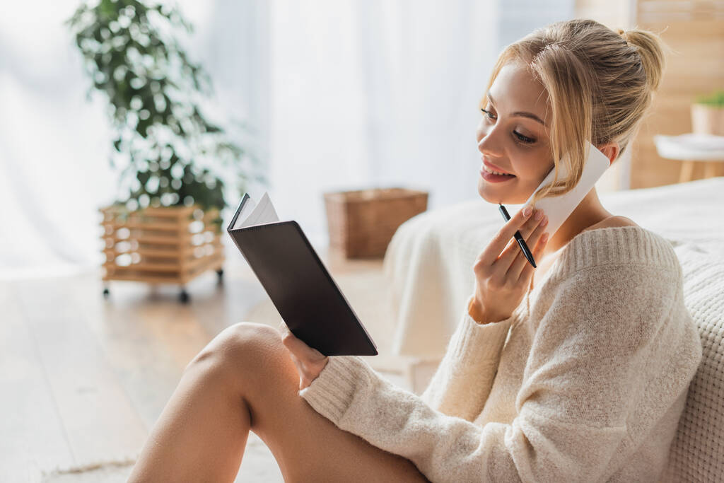 cheerful woman in sweater and socks talking on smartphone and looking at notebook  - Photo, Image
