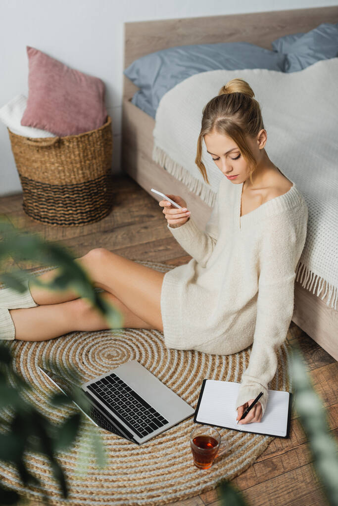 mulher bonita em suéter tomando notas perto do laptop e segurando smartphone enquanto sentado perto da cama  - Foto, Imagem