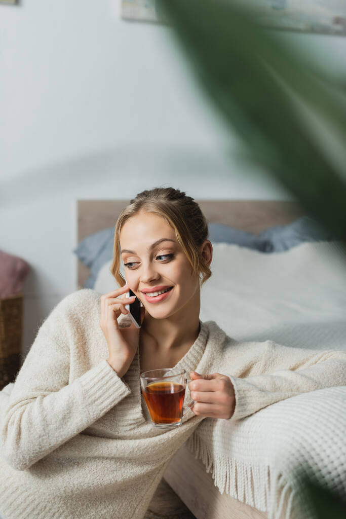 Zufriedene Frau spricht auf Smartphone und hält Glas Tee im Schlafzimmer  - Foto, Bild