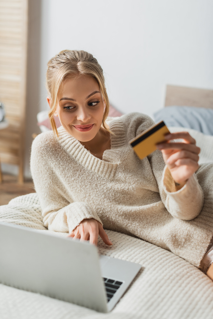 femme gaie tenant carte de crédit et en utilisant un ordinateur portable tout en faisant des achats en ligne dans la chambre  - Photo, image