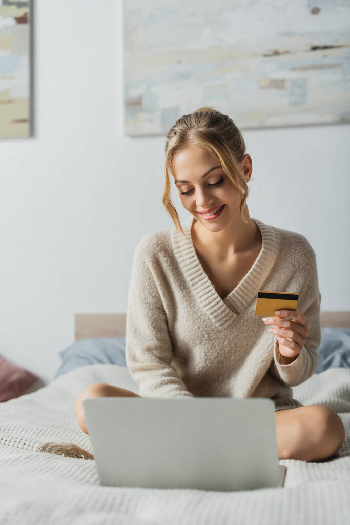Yatak odasında online alışveriş yaparken bilgisayarın yanında kredi kartı tutan tatmin olmuş bir kadın.  - Fotoğraf, Görsel
