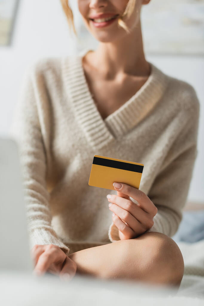 vue recadrée d'une femme souriante tenant une carte de crédit près d'un ordinateur portable et assise sur son lit - Photo, image