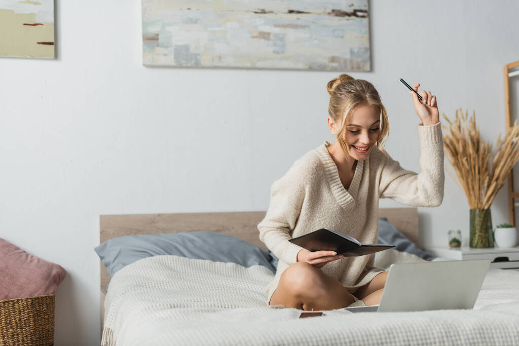 vidám fiatal nő gazdaság notebook és toll, miközben tanul online a hálószobában  - Fotó, kép
