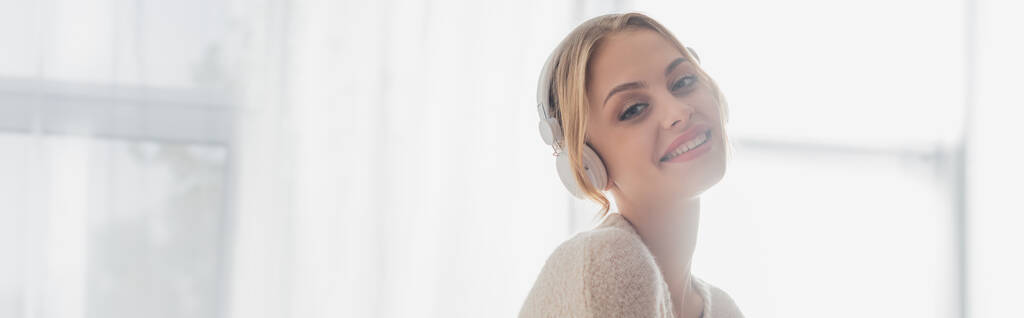 overjoyed young woman listening music in wireless headphones while smiling at home, banner  - Photo, Image