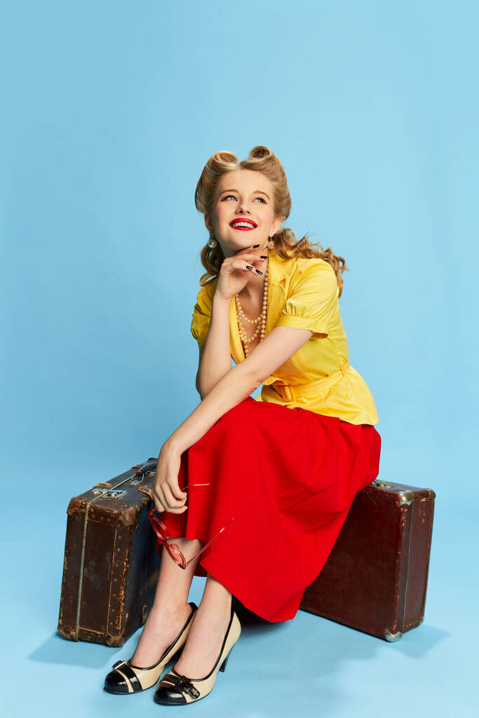 Álmodozó, boldog tekintet. Gyönyörű fiatal lány sárga blúzban és piros szoknyában ül a bőröndön kék stúdió háttér mellett. Fogalom retro divat, szépség, utazás, 50-es, 60-as évek. Pin-up stílus - Fotó, kép