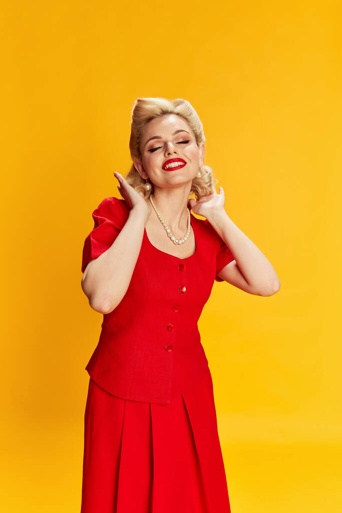 Positive femininity. Beautiful young girl with stylish hairstyle in red dress posing against yellow studio background. Concept of retro fashion, beauty, 50s, 60s. Pin-up style - Photo, Image