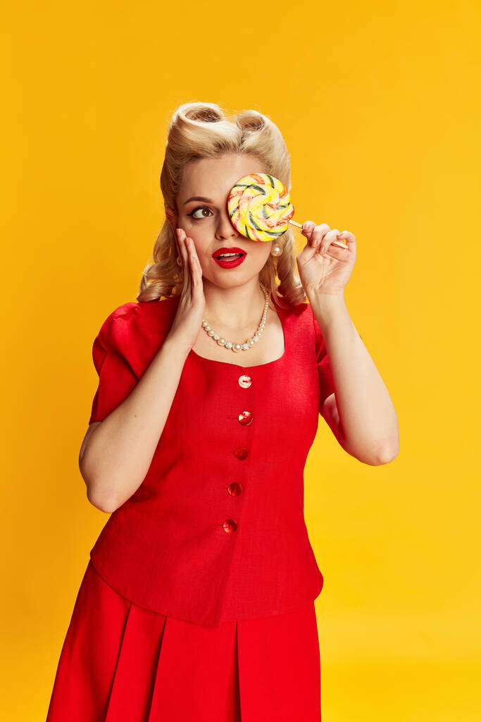 Milovník bonbónů. Portrét krásné mladé blondýny se stylovým účesem v červeném obleku pózující proti žlutému pozadí studia. Koncept retro módy, krásy, 50. a 60. let. Pin-up styl - Fotografie, Obrázek