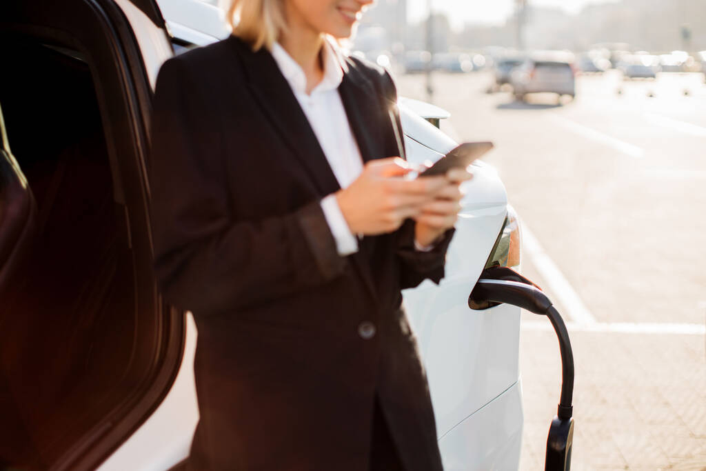 Close up de mulher caucasiana agradável em roupas formais com smartphone moderno em mãos de pé perto de carregar carro electro ao ar livre. Fundo borrado da área urbana. - Foto, Imagem