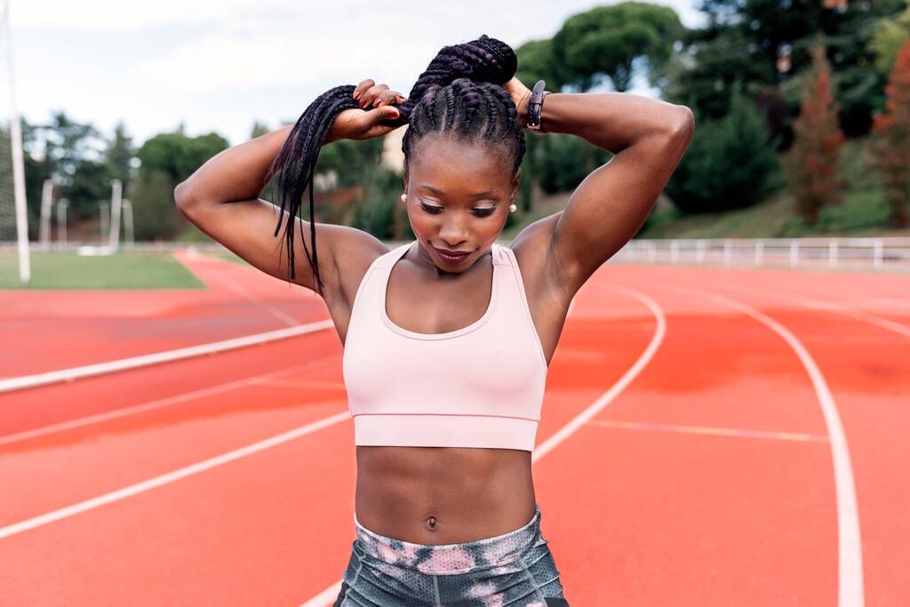 Archivbild einer afroamerikanischen Sprinterin, die auf einer Leichtathletikbahn steht und ihre geflochtenen Haare mit der Hand berührt. Freizeit - Foto, Bild