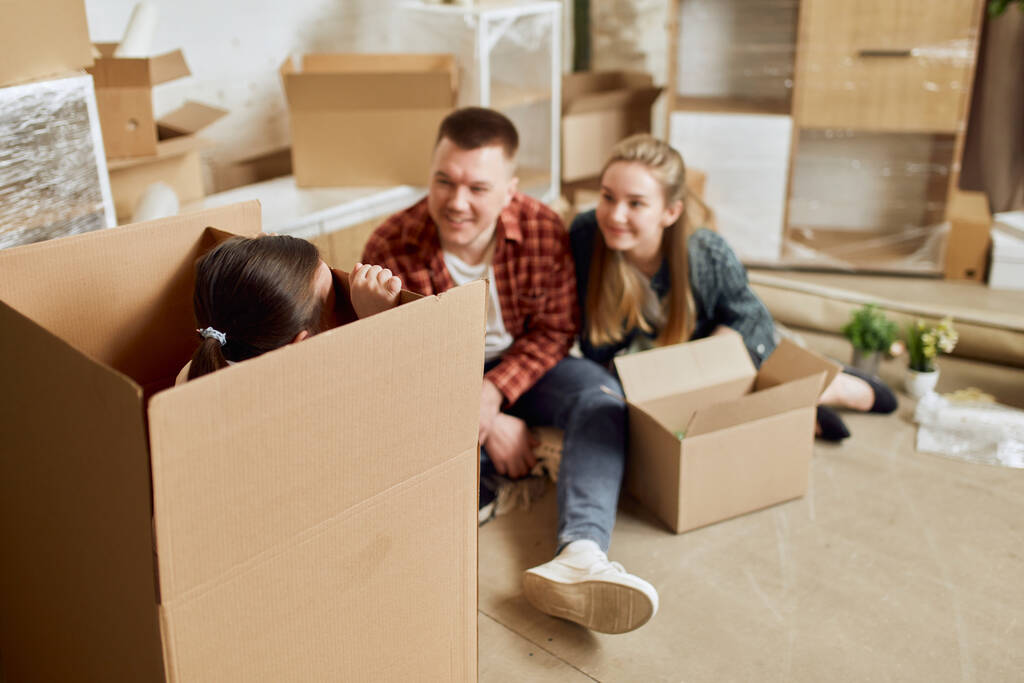Little girl hiding into box, playing with parents. Young happy family, man, woman and kid moving into new apartment with many cardboard boxes. Concept of moving houses, real estate, family, new life - Photo, Image