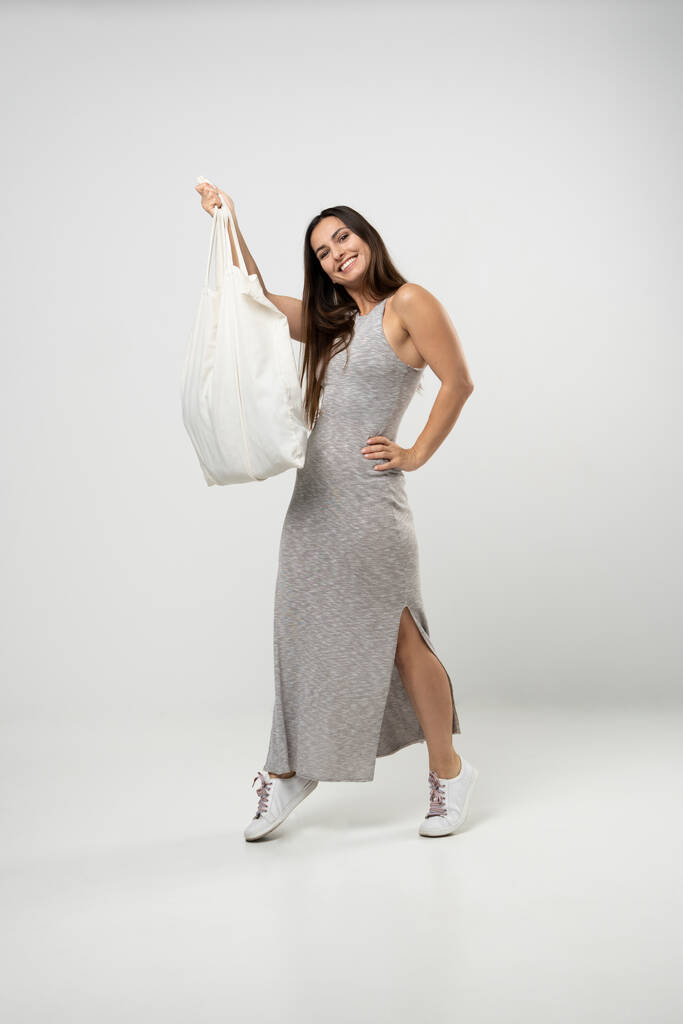 Brünette Frau im grauen Kleid, Baumwolltasche mit Gemüse, Produkte im weißen Raum. Umweltfreundliche Einkaufstüten. Null Abfall, plastikfreies Konzept - Foto, Bild