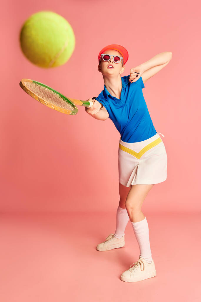 Piłka uderzeniowa. Portret pięknej młodej dziewczyny w sportowej odzieży pozującej z rakietą tenisową na różowym tle. Pojęcie młodości, sportu, mody, stylu życia, emocji, wyrazu twarzy. Ogłoszenie - Zdjęcie, obraz