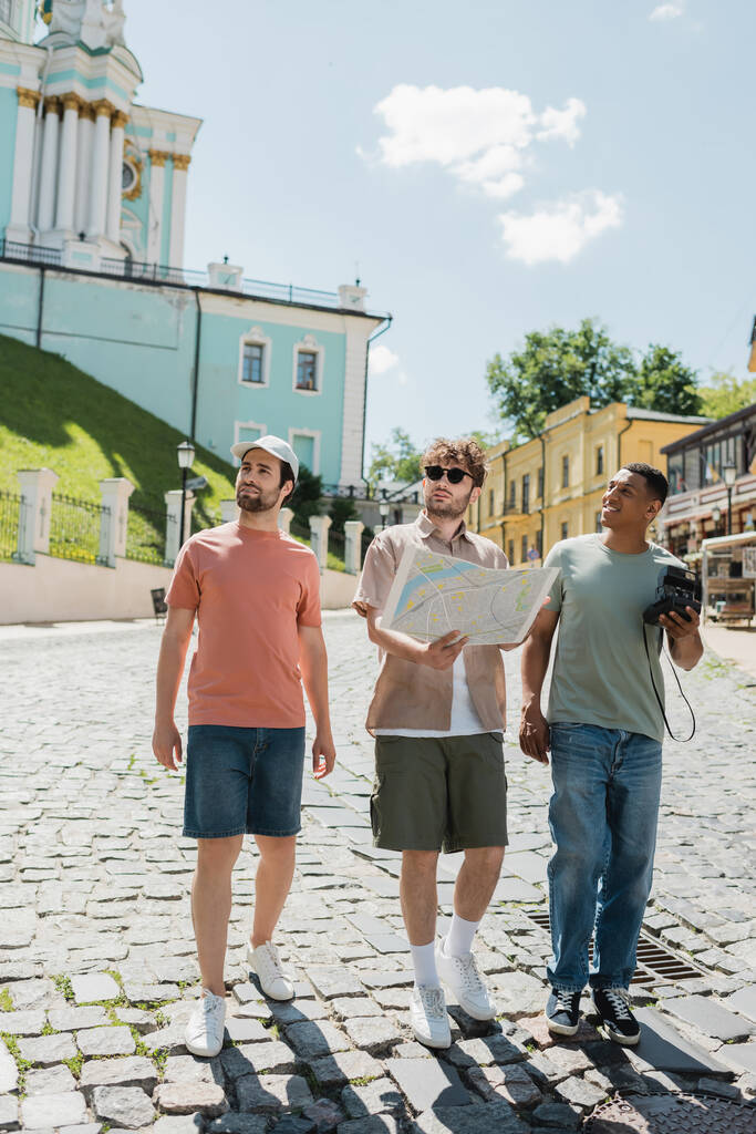 longitud completa de los turistas interracial con cámara vintage y mapa de la ciudad caminando con guía en el descenso de Andrews en Kiev - Foto, Imagen