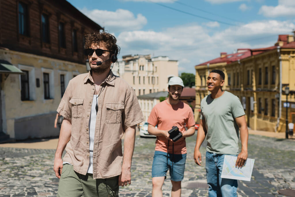 guida turistica in occhiali da sole e auricolare a piedi vicino a turisti multiculturali offuscati sulla discesa Andrews a Kiev - Foto, immagini