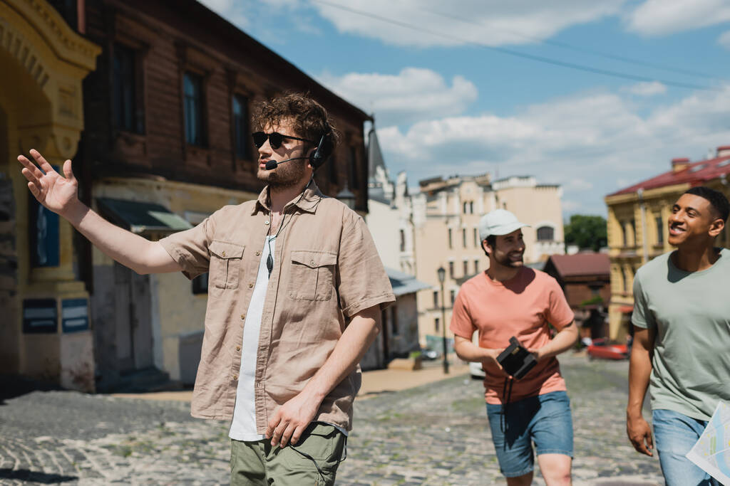 giovane guida in auricolare e occhiali da sole che punta con mano durante l'escursione con uomini interrazziali sul quartiere Podil a Kiev - Foto, immagini