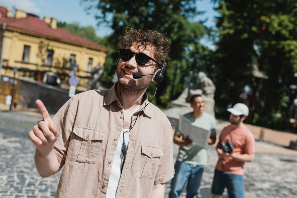 junger und unbeschwerter Führer mit Sonnenbrille und Headset, der mit dem Finger in der Nähe verschwommener interrassischer Touristen im Kiewer Stadtteil Podil zeigt - Foto, Bild