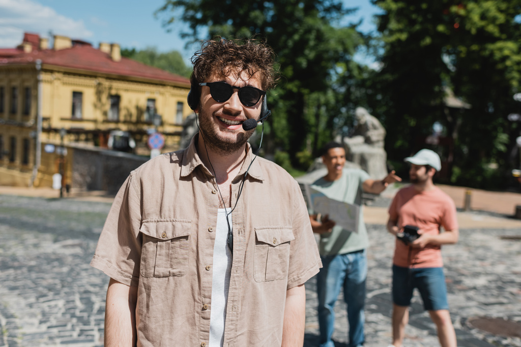 ξέγνοιαστος ξεναγός σε ακουστικά και γυαλιά ηλίου χαμογελώντας κοντά σε θολή πολυεθνική ταξιδιώτες για Andrews καταγωγή στο Κίεβο - Φωτογραφία, εικόνα