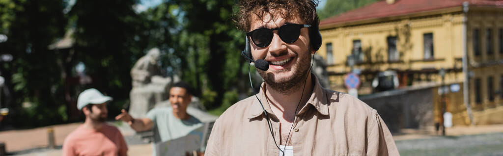 retrato de jovem guia turístico em fone de ouvido e óculos de sol perto turvo turistas inter-raciais em Andrews descida em Kiev, banner - Foto, Imagem