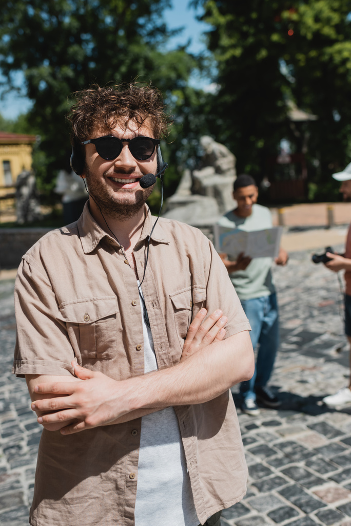 νεαρός και χαρούμενος ξεναγός σε γυαλιά ηλίου και ακουστικά που στέκονται με σταυρωμένα χέρια στην κατάβαση Andrews στο Κίεβο κατά τη διάρκεια της καλοκαιρινής ημέρας - Φωτογραφία, εικόνα