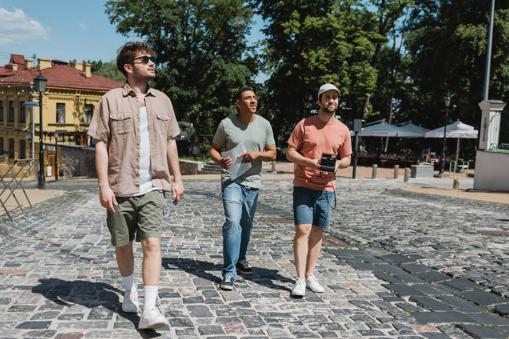 полная длина экскурсовода и мультикультурных путешественников с картой и винтажной камерой, гуляющих по тротуару Эндрюса спуска в Киеве - Фото, изображение